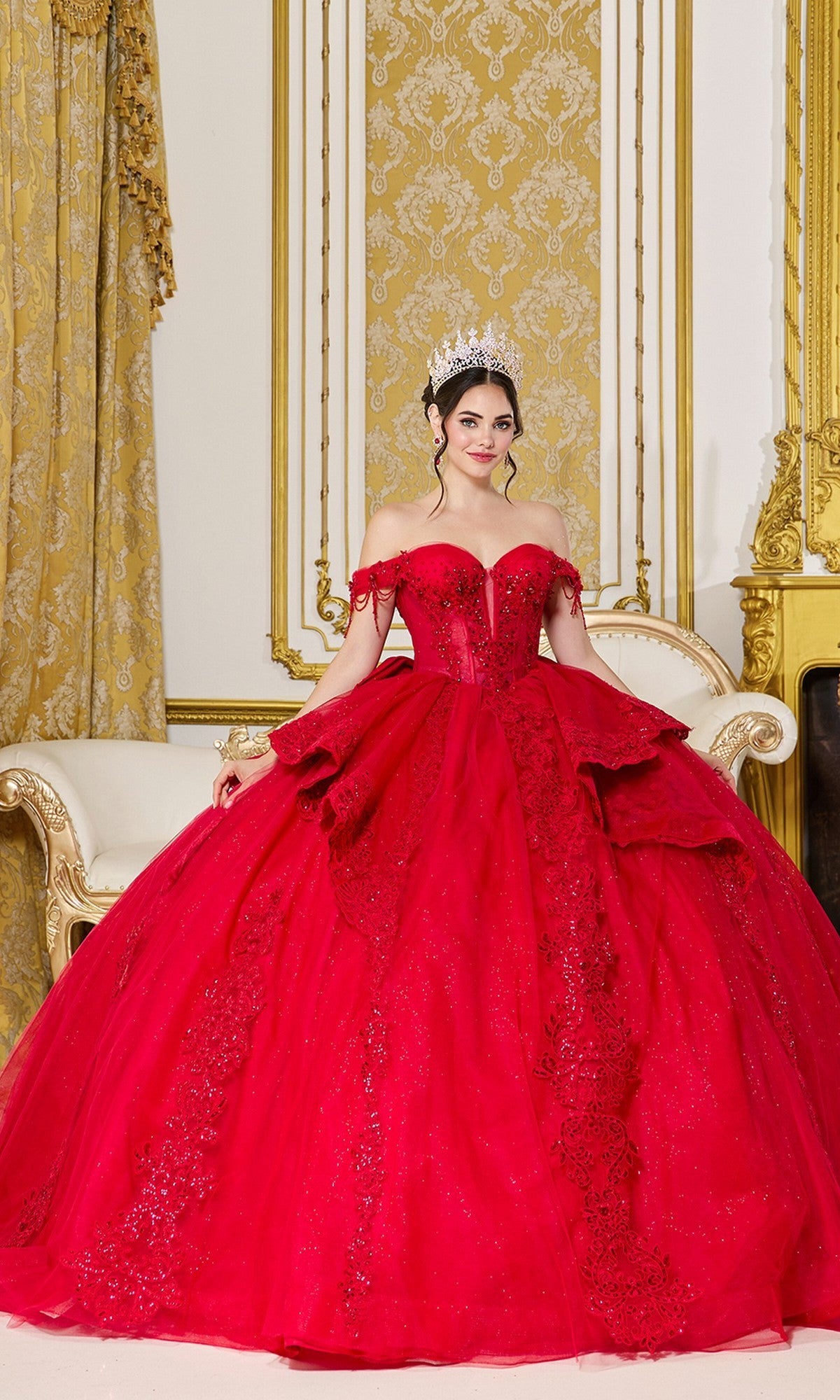A woman in the Quinceanera Dress 5108 By Dancing Queen, a sparkling red gown with a corset bodice, and a tiara stands in an ornate room with gold and white decor.